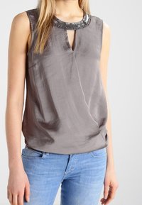 Mouwloze grijze satijnen blouse met een geperleerde halslijn, gedragen met lichtblauwe jeans door een staand persoon.