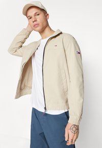 Bežani lagani jaknu s crnim patentnim zatvaračem, elastičnim manžetama i Tommy Hilfiger logotipom na prsima i rukavu. Nosena preko bijele majice.
