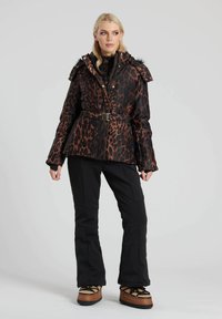 Winterjacke im Leopardenmuster mit einer mit Kunstfell besetzten Kapuze, tailliertem Gürtel und Reißverschlusstaschen, kombiniert mit schwarzen Skihosen und braunen Stiefeln.