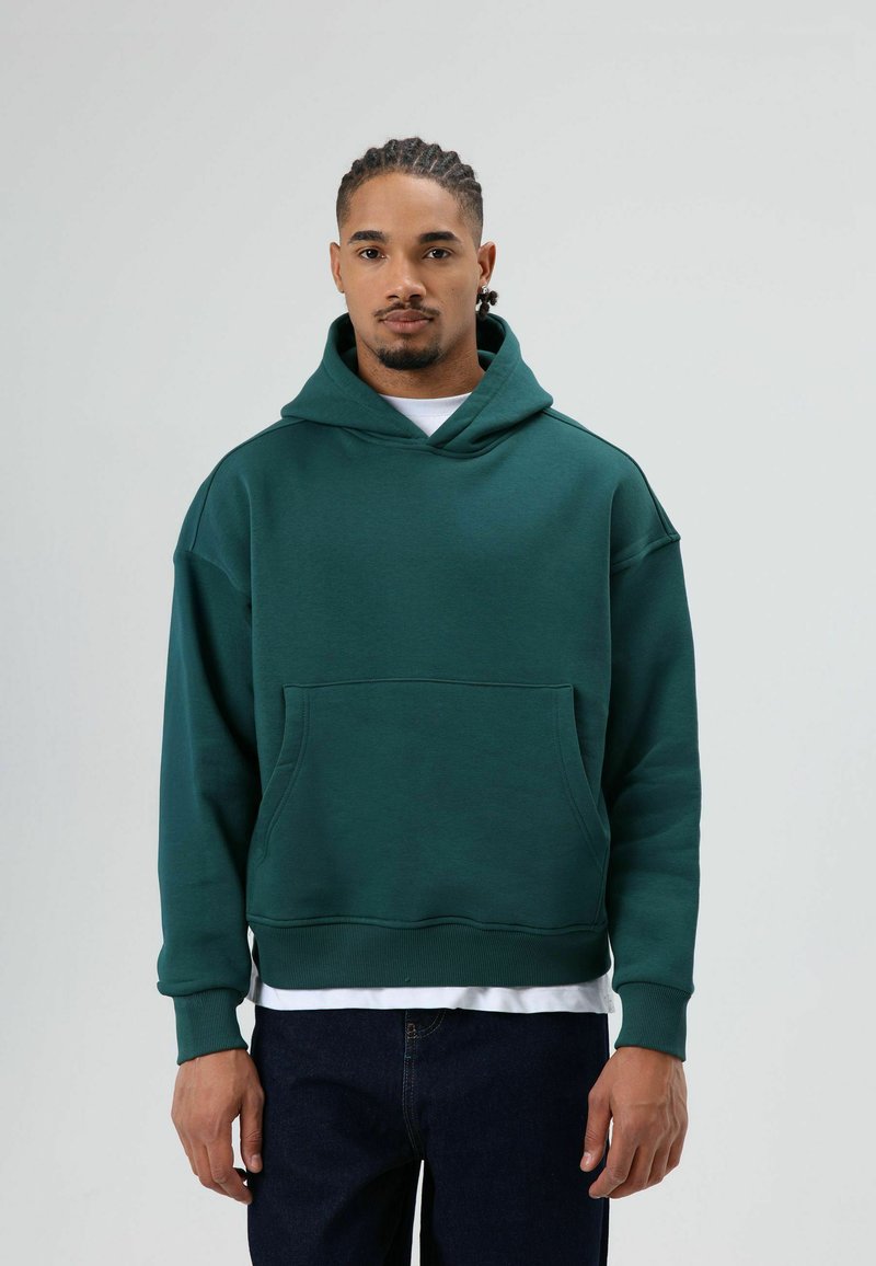 Sweat à capuche en coton vert avec une poche frontale, une capuche et des poignets côtelés. Porté sur une chemise blanche, associé à un jean en denim foncé.