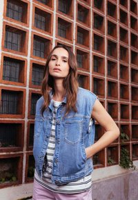 Denim vest met een mouwloos ontwerp, lichtblauwe kleur, twee voorzakken, gedragen over een gestreepte shirt. Achtergrond met een gestructureerde bakstenen muur.