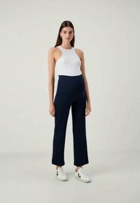 Marineblau hochgeschnittene Hose mit geradem Bein, kombiniert mit einem weißen ärmellosen Oberteil. Das Modell trägt weiße Sneakers und zeigt einen lässigen Look.