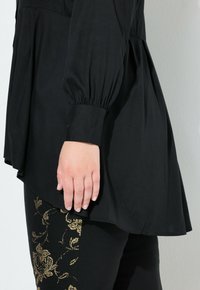 Schwarze Bluse mit Ballonärmeln und einem Peplum-Saum, kombiniert mit schwarzen Leggings, die goldene Blumenstickereien an der Seite aufweisen.