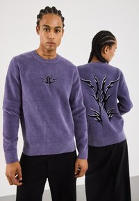 Paarse gebreide trui met ronde hals, met een zwart geborduurd ontwerp op de voor- en achterkant, ribbelmanchetten en -zoom. Draag met zwarte broek.