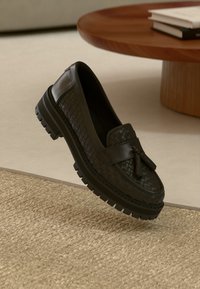 Black woven leather loafer with a chunky black rubber sole and tassel detail, suspended above a textured beige surface.
