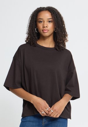 Kvinde med krøllet hår iført en løs sort T-shirt og sølvringe står mod en ensfarvet lys baggrund og kigger fremad.