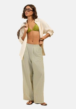 Femme portant une chemise beige oversize par-dessus un haut de bikini vert, un pantalon large rayé, des sandales, des lunettes de soleil, des bracelets en bois et portant un sac tressé.