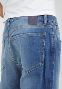 Modré džíny z denimu s mírně vybledlým vzhledem, vybavené koženým štítkem značky, klasickými pěti kapsami a kontrastním šitím.
