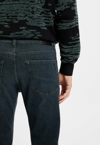 Hombre vistiendo vaqueros azul oscuro y un suéter negro con patrón verde, mostrado desde la cintura hacia abajo y ligeramente girado de espaldas.