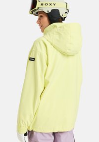 Casaco amarelo claro com capuz, punhos elásticos e um logo preto da Roxy na manga. Usado com uma luva branca e um capacete de esqui.