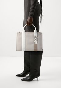 Sac fourre-tout en toile beige avec des accents blancs et taupe, doté d'une fermeture éclair en haut et d'un logo "DKNY" brodé sur le devant.