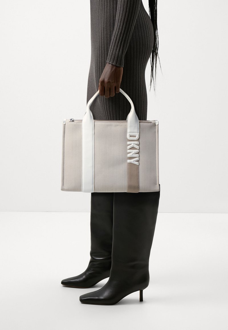 Sac fourre-tout en toile beige avec des accents blancs et taupe, doté d'une fermeture éclair en haut et d'un logo "DKNY" brodé sur le devant.