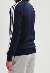 Giacca zip-up blu navy con strisce bianche sulle maniche, realizzata in tessuto liscio, con colletto alto e orlo a costine.