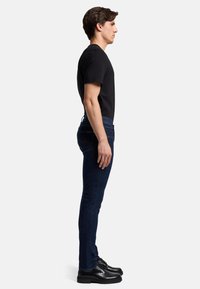 Jeans aderenti in denim blu scuro con una finitura liscia, abbinati a una T-shirt nera aderente e stivali neri stringati, visti di profilo.