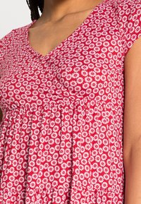 Mujer vistiendo un vestido rojo con un patrón floral de margaritas blancas y un escote en V, mostrando la parte superior del torso y el brazo derecho.