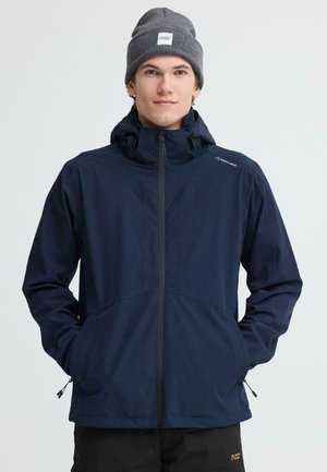 Noor mees, kes kannab tumedat sinist kapuutsiga lukuga jope ja halli mütsi, seisab kätega taskutes ühevärvilise tausta ees.