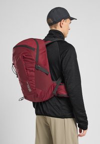 Roter Rucksack mit einem eleganten Design, der graue Akzente, mehrere Fächer und verstellbare Träger aufweist. Wird über einem schwarzen strukturierten Stoff getragen.