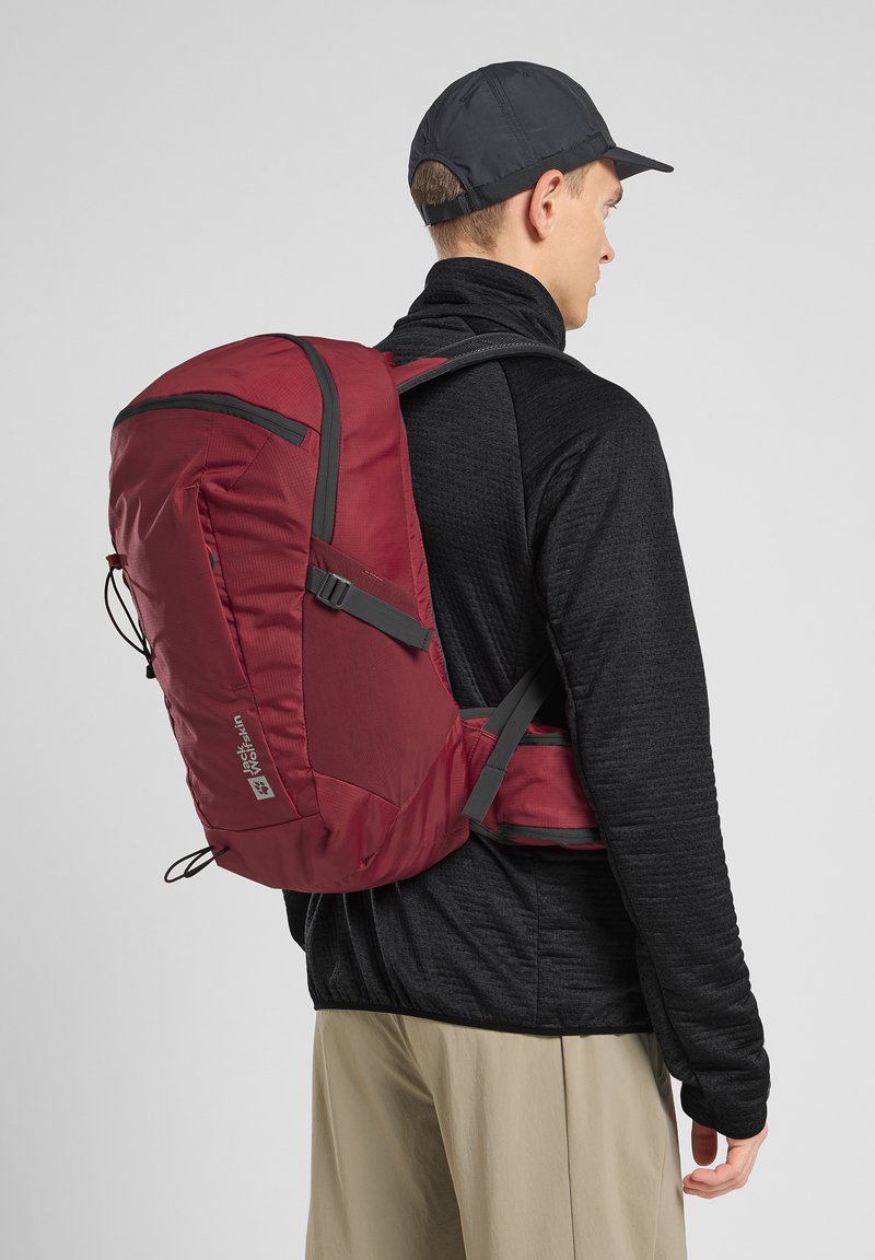 Roter Rucksack mit einem eleganten Design, der graue Akzente, mehrere Fächer und verstellbare Träger aufweist. Wird über einem schwarzen strukturierten Stoff getragen.