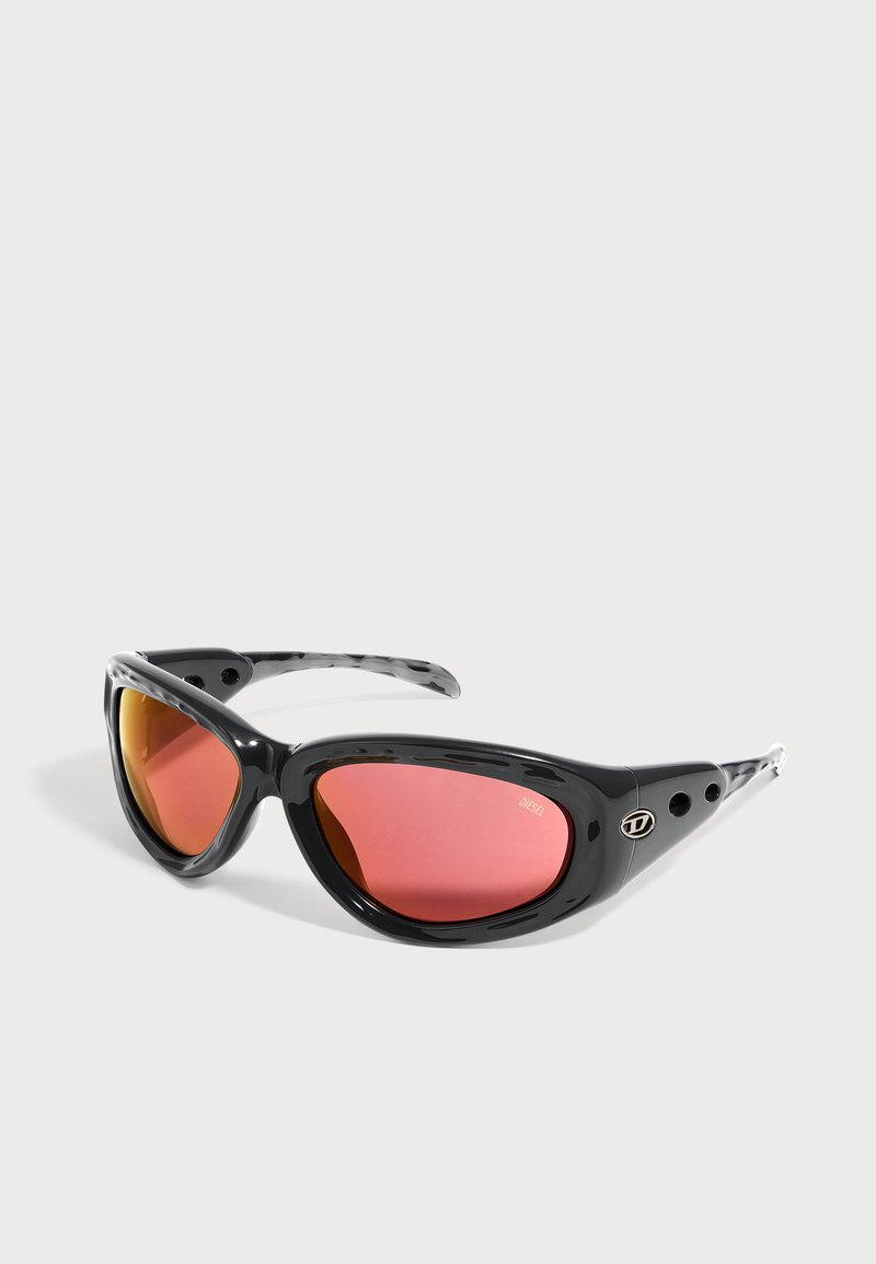 Lunettes de soleil enveloppantes noires avec des verres réfléchissants rouges et de petits trous de ventilation sur les branches, inclinées sur fond blanc.
