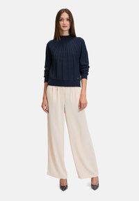 Granatowy sweter dzianinowy z wysokim prążkowanym kołnierzem i wzorem w warkocze, zestawiony z jasnobeżowymi spodniami o szerokich nogawkach i czarnymi szpiczastymi szpilkami.