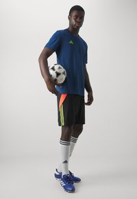 Tricou athletic albastru, pantaloni scurți negri cu accente portocalii neon și galbene, șosete albe cu dungi negre �și ghete de fotbal albastre. Ține o minge de fotbal.