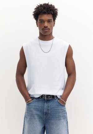 Fiatal férfi rövid, göndör hajjal, ujjatlan fehér ingben, ezüst lánc nyaklánccal, ezüst karkötővel, fekete övvel és kék farmernadrággal, zsebre tett kézzel.