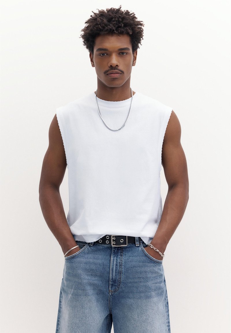 Mlad moški s kratkimi kodrastimi lasmi, ki nosi belo majico brez rokavov, srebrno verižico, srebrno zapestnico, črni pas in modre kavbojke, z rokami v žepih.