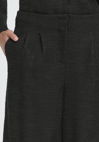 Pantaloni neri con trama a tessuto, fronte plissettato e tasche laterali, realizzati in tessuto pesante.