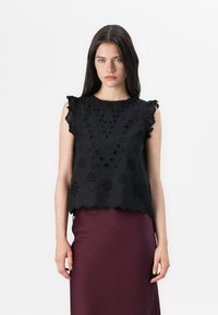 Model, oblečena v črno vezeno bluzo z nabranimi rokavi in maroon krilu, stoji na belem ozadju.