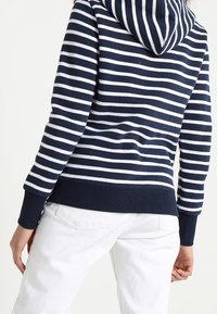 Sudadera a rayas en azul marino y blanco con capucha ajustable, puños y dobladillo acanalados, combinada con pantalones blancos. Material de algodón, corte casual.
