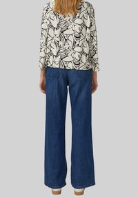 Florale Bluse mit langen Ärmeln in Schwarz-Weiß-Mustern, kombiniert mit locker sitzenden, dunkelblauen Jeans mit weitem Bein und hellen Schuhen.