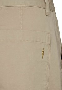 Bēša kravas šorti ar aizmugurējo kabatu un zelta zibens zīmējumu. Audums ir gluds ar izturīgu struktūru un kvalitatīvu šuvumu.