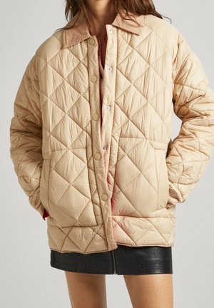 Frau trägt eine beige gesteppte Jacke mit großem Kragen und Druckknöpfen, kombiniert mit einem schwarzen Minirock mit Reißverschluss vorne. Hände in den Taschen.