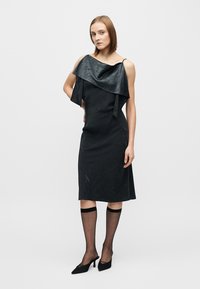 Robe noire avec un col asymétrique drapé et une silhouette ajustée. Portée avec des bas noirs transparents et des talons pointus. Texture lisse.