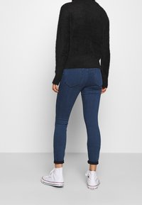 Persona vistiendo un suéter negro de peluche, jeans ajustados oscuros enrollados y zapatillas Converse de plataforma blancas, de pie contra un fondo simple.