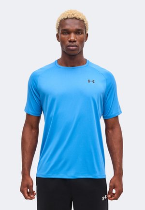 Mann mit kurzen lockigen blonden Haaren, der ein hellblaues kurzärmeliges Sportshirt und schwarze Hosen vor einem einfarbigen Hintergrund trägt.