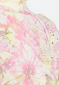 Blouse transparente à motif floral rose et jaune, avec détails brodés et découpes œillets, col montant.