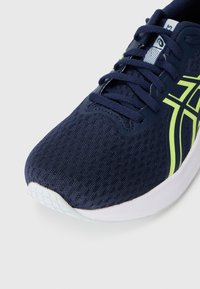 Baskets de sport bleu marine avec une tige en maille respirante, des accents vert fluo, et une semelle blanche avec un motif de grip texturé.