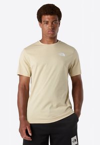 Beżowy t-shirt z krótkim rękawem wykonany z gładkiej tkaniny, z białym logo po lewej stronie klatki piersiowej. Noszony z czarnymi szortami z logo.