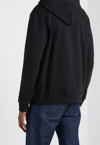 Black hooded sweatshirt with a smooth texture, ribbed cuffs, and hem. The back is plain with a central seam, paired with dark blue jeans.