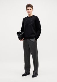 Il maglione nero a collo rotondo presenta un dettaglio del logo in rilievo. Abbinato a pantaloni grigi a quadri e scarpe oxford nere. Tiene una pochette nera.
