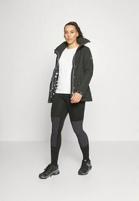 Fekete vízálló dzseki magas gallérral, ezüst színű kiegészítőkkel és pöttyös béléssel, fehér ing és szürke részletekkel díszített fekete leggings fölött viselve.