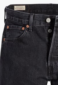 Vue de face d'un jean denim noir Levi's premium montrant la braguette à boutons, les passants de ceinture, la poche avant avec rivets en cuivre et l'étiquette Levi's à l'intérieur de la ceinture.
