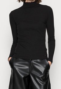 Vrouw in een aansluitend zwart geribbeld longsleeve met een gerimpelde halslijn en losse zwarte faux leren broek met handen in de zakken.