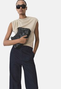 Svart vävd handväska hålls mot en ljusbeige topp, kombinerad med mörkblå vida jeans och stora svarta solglasögon.