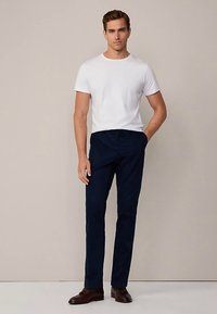 Weißes Baumwoll-T-Shirt unter dunkelblauen Hosen, kombiniert mit braunen Lederschuhen. Das Outfit besticht durch eine lässige Passform und eine glatte Stoffstruktur.