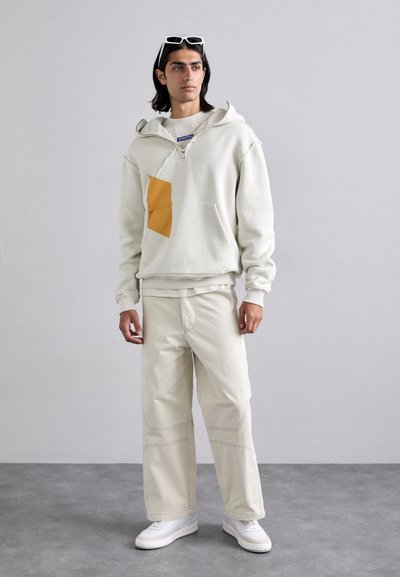 Un hoodie gris clair avec une poche géométrique jaune, associé à un pantalon large crème et des baskets blanches. Tissu doux avec une coupe décontractée.