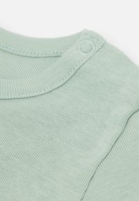 Jasnozielona bawełniana koszula z prążkowaną strukturą, zaokrąglonym dekoltem i zapięciem na napy na ramieniu, ułatwiającym ubieranie.