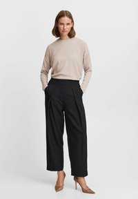 Beige ribbed long-sleeve top paired with high-waisted black pleated trousers and brown pointed-toe heels, standing on a neutral background.
