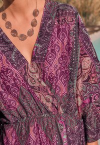 Rochie tip wrap cu model paisley mov, decolteu în V, talie strânsă și mâneci scurte și vaporoase. Colier argintiu cu frunză accentuează linia gâtului.